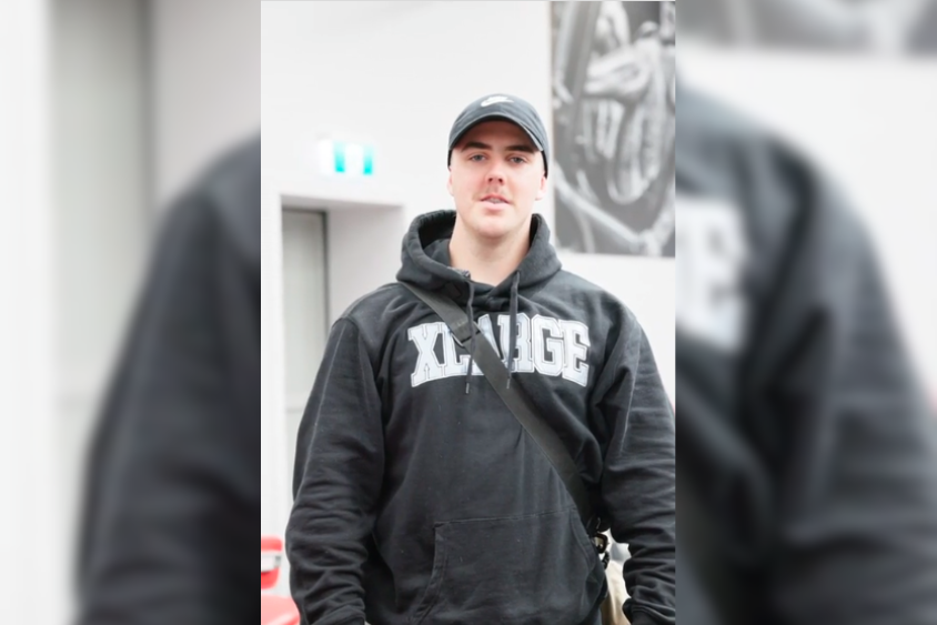 A young man stands confidently in a black hoodie with the word "XLARGE" printed on it, wearing a cap. He is in a gym setting, showcasing a friendly demeanour that reflects the welcoming community at Genesis Ballarat, where he has been a member for about 11 months.