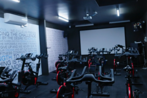 An indoor cycling studio at Genesis Ballarat features several stationary bikes arranged in rows. The walls are adorned with motivational text, and a large projection screen is positioned at the front of the room. The space is brightly lit, creating an inviting atmosphere for group cycling classes, emphasising the gym's commitment to providing top-quality equipment and training options.