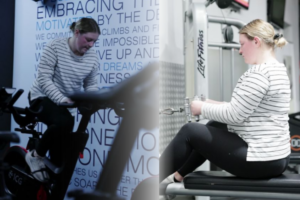 A young woman engages in a workout at Genesis Ballarat, alternating between a stationary bike and a strength training machine.