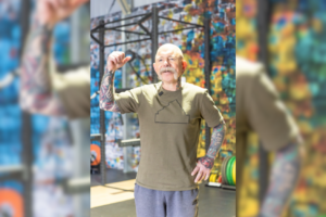 Mitchell stands happily in a gym at Genesis Ballarat, smiling and raising his hand. He has tattoos and wears a green shirt and grey pants. The colourful wall behind him shows the lively atmosphere of the gym.
