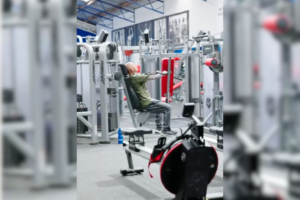 Mitchell is working out in a gym at Genesis Ballarat. He is seated on a weight machine, focusing on his exercise. The gym has various equipment in the background, showing a lively and well-equipped space. 