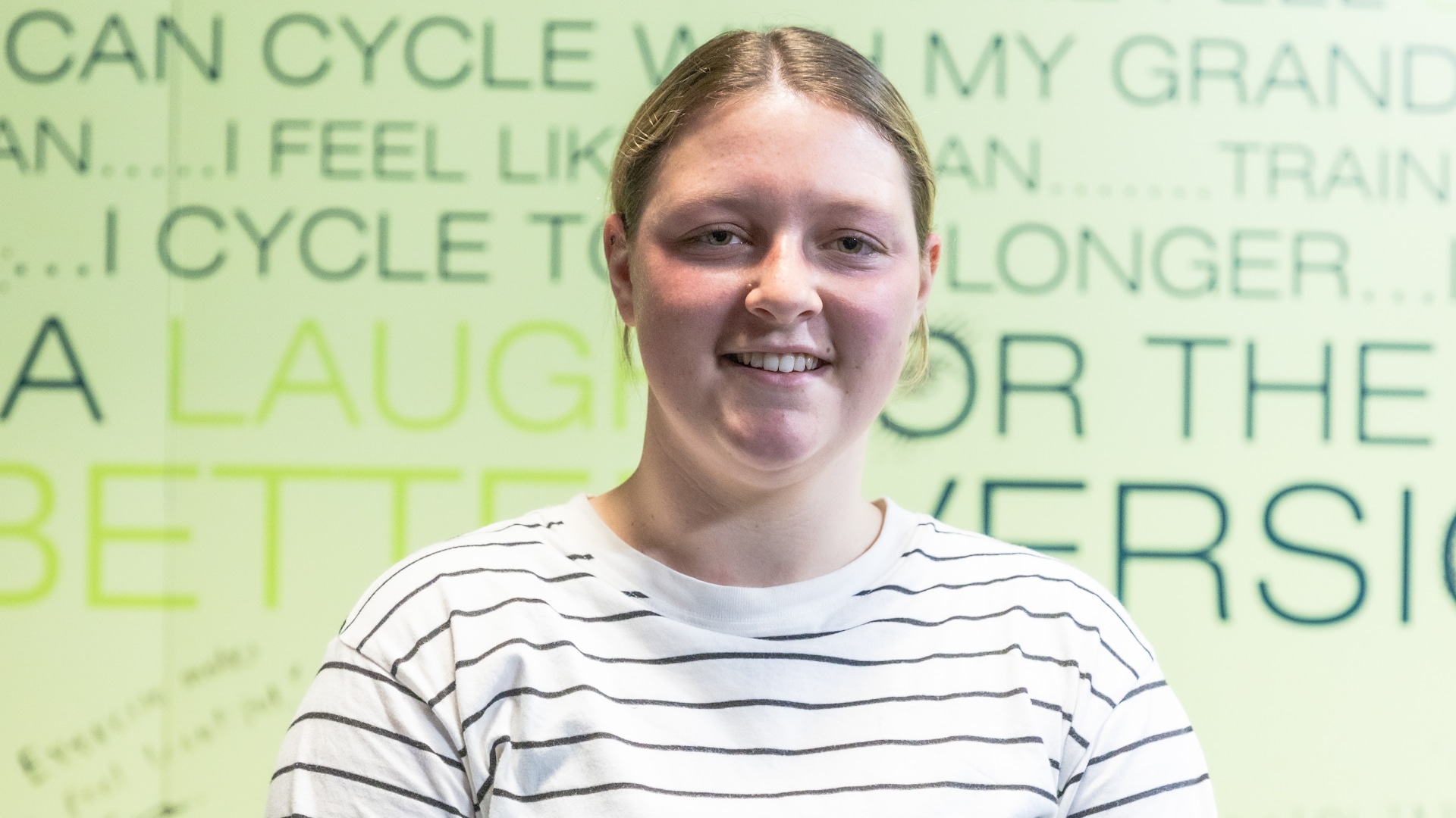 A smiling young woman named Taliah stands in front of a bright green wall with motivational text, wearing a striped shirt. The image features the text: "I CAME FOR THE GROUP CLASSES & FELL IN LOVE WITH STRENGTH TRAINING - TALIAH," highlighting her fitness journey at Genesis Ballarat.