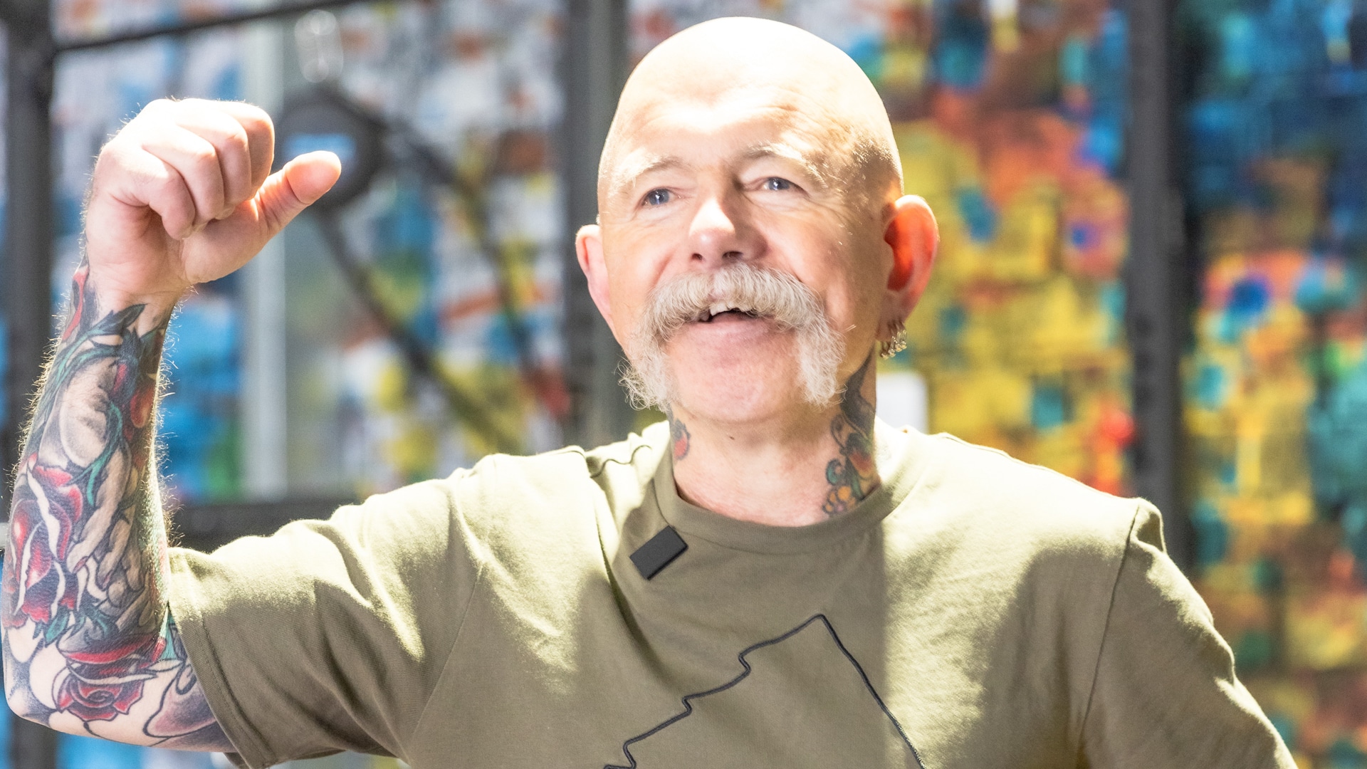 Mitchell, an enthusiastic older man with a distinctive moustache and tattoos, is joyfully raising his fist in celebration. He is wearing a casual shirt and has a bright smile, embodying his love for fitness. The image includes the text: "I JUST LOVE EVERY INCH OF THE GYM - MITCHELL," highlighting his passion for Genesis Ballarat and his 40-year fitness journey.